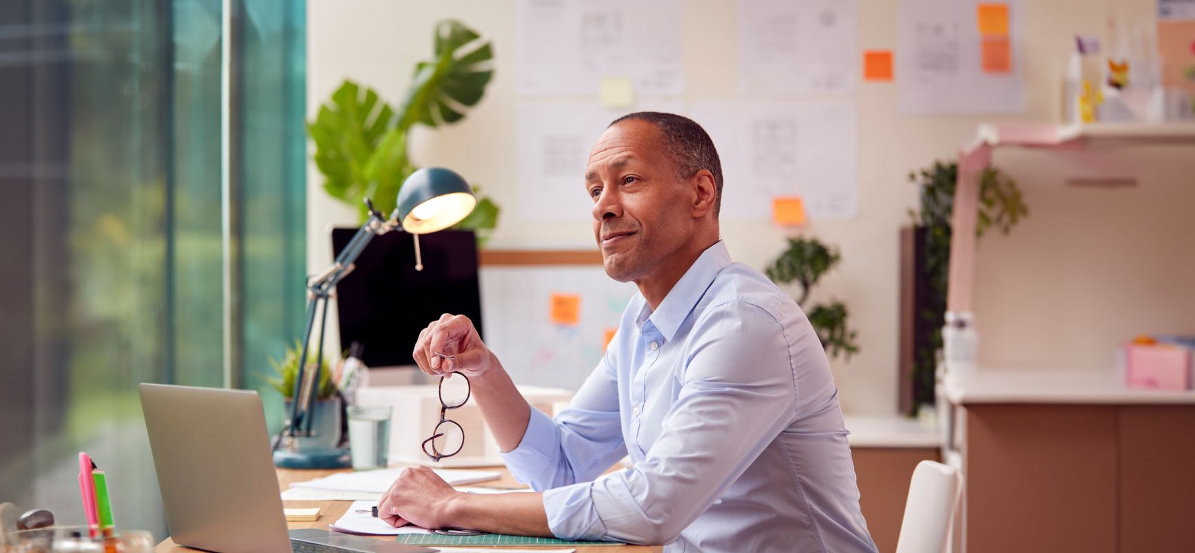 Man sitting at desk holding reading glasses and thinking